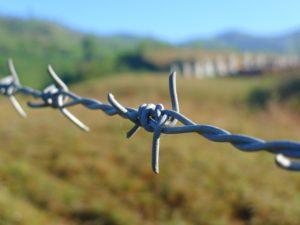 Picture of barbed wire up close with grassland, hills and blue sky out of focus in the background.